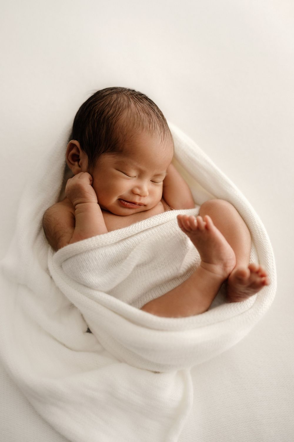 A peaceful newborn baby wrapped in soft white blanket sleeps contentedly during professional portrait session.