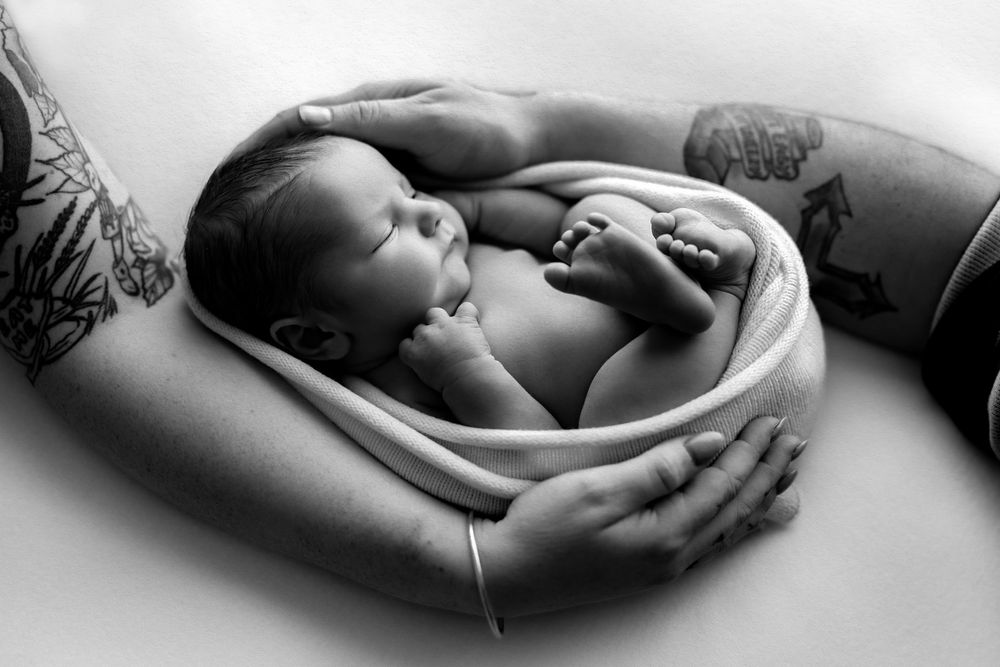 Black and white photo of a sleeping newborn baby cradled in tattooed parents hands.