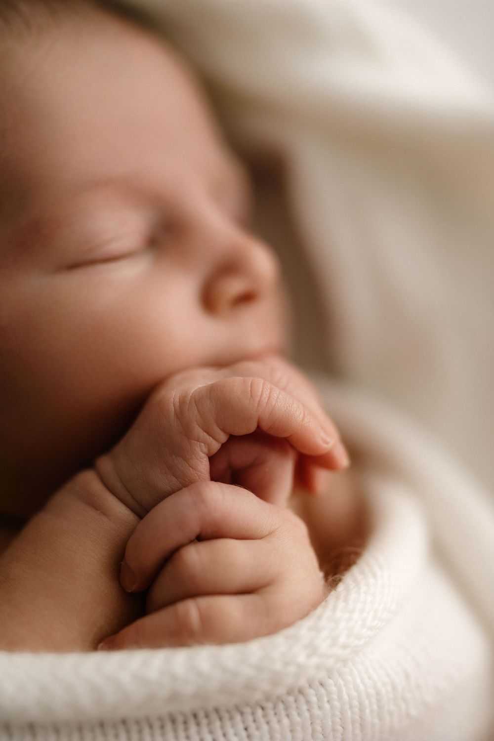 Peaceful newborn baby sleeping wrapped in soft white knit blanket with tiny hands tucked under chin.