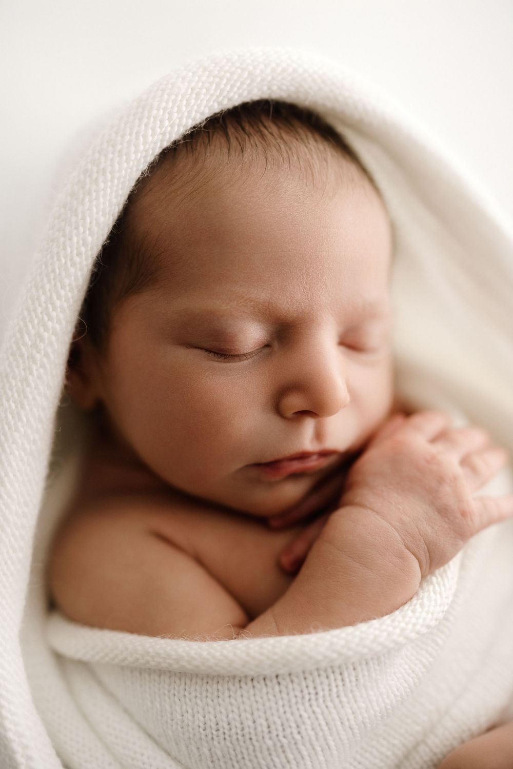 Peaceful newborn baby sleeping wrapped in a soft white knit blanket with hands tucked under chin.