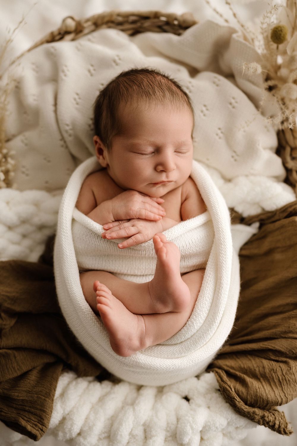 Peaceful newborn baby sleeping in a wicker basket, wrapped in a white knit blanket with tiny feet peeking out.