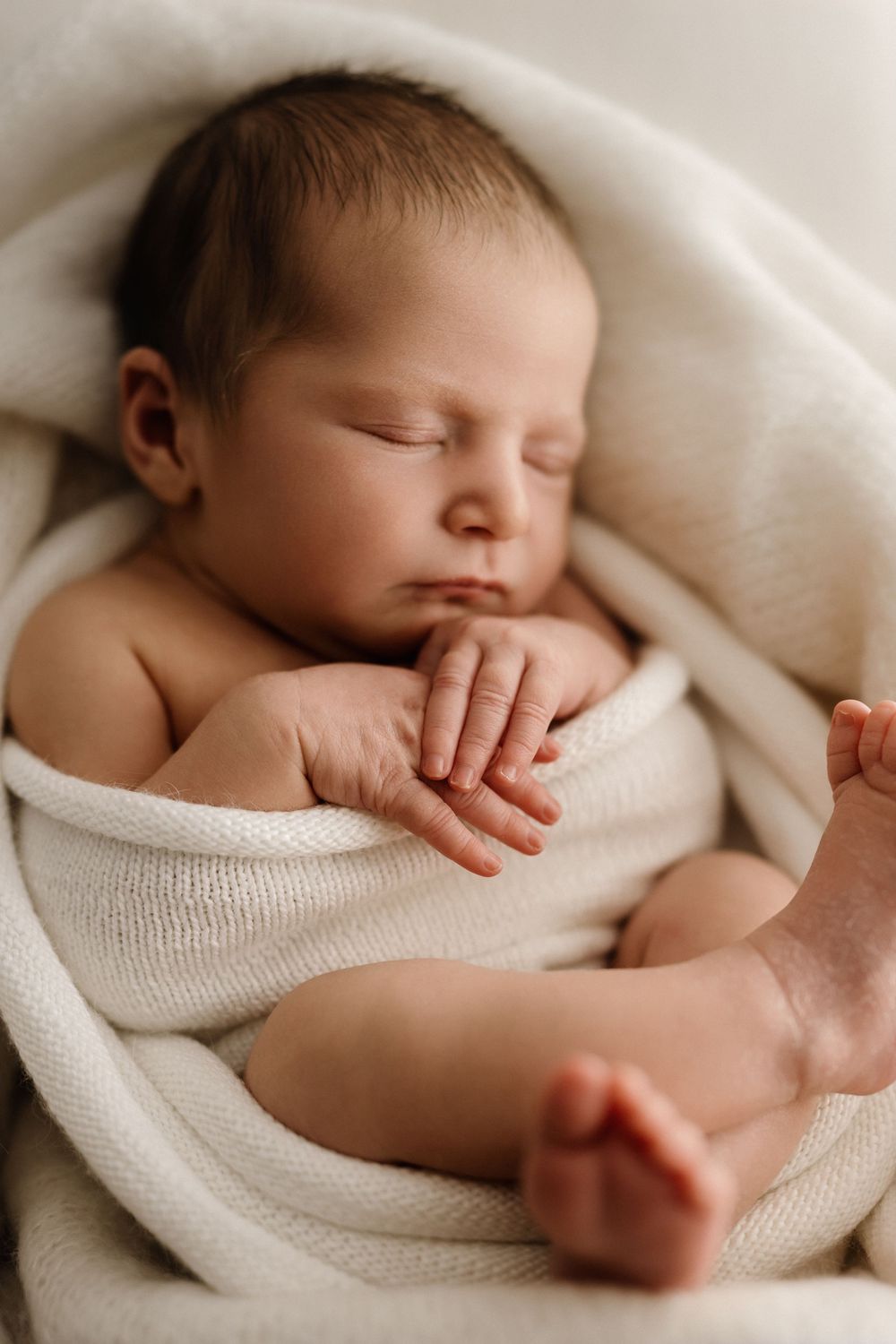 Peaceful newborn baby sleeping wrapped in a soft white knit blanket with tiny hands and feet visible.
