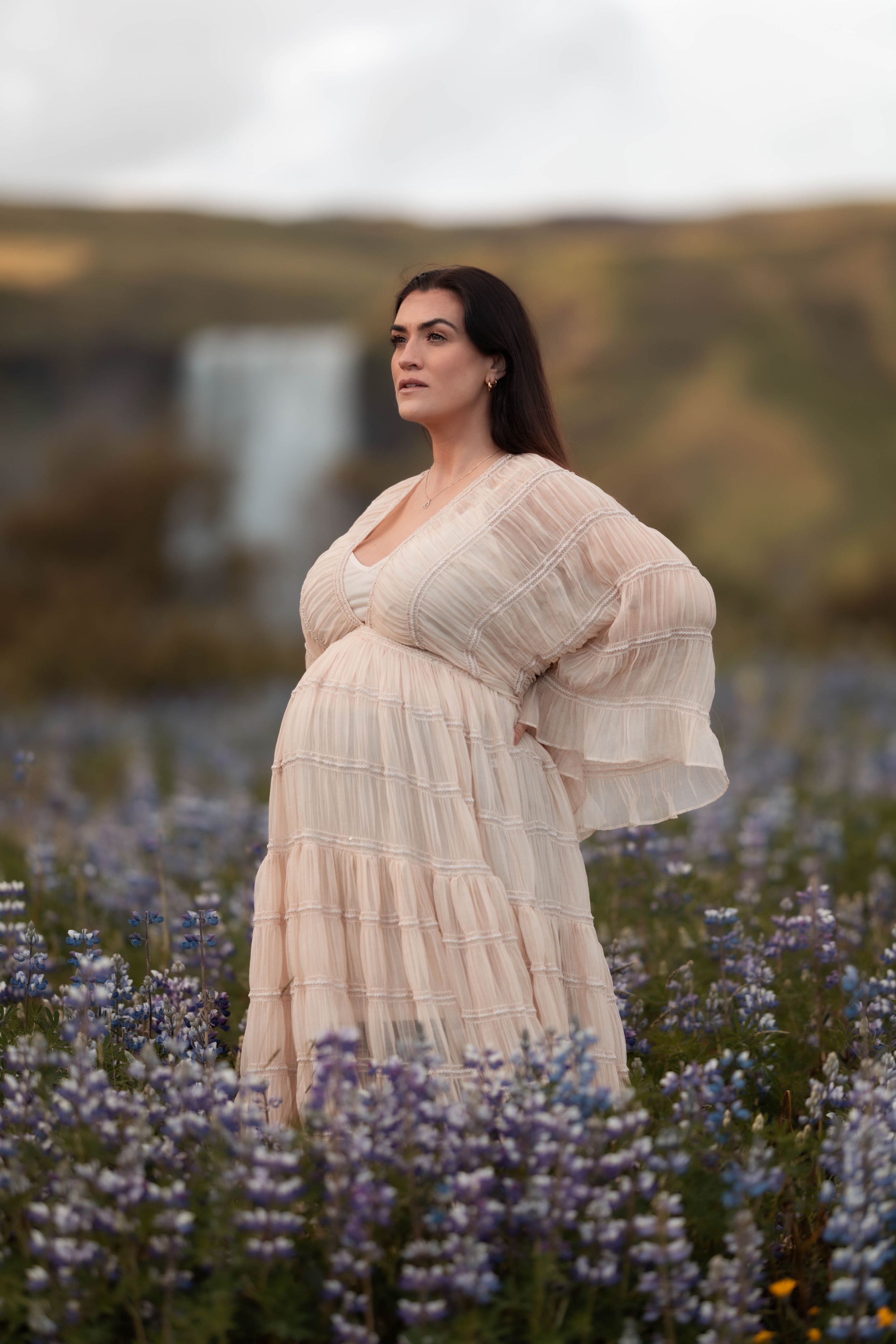 Pregnant woman in blush ruffled dress stands in purple lupine flower field under cloudy sky.