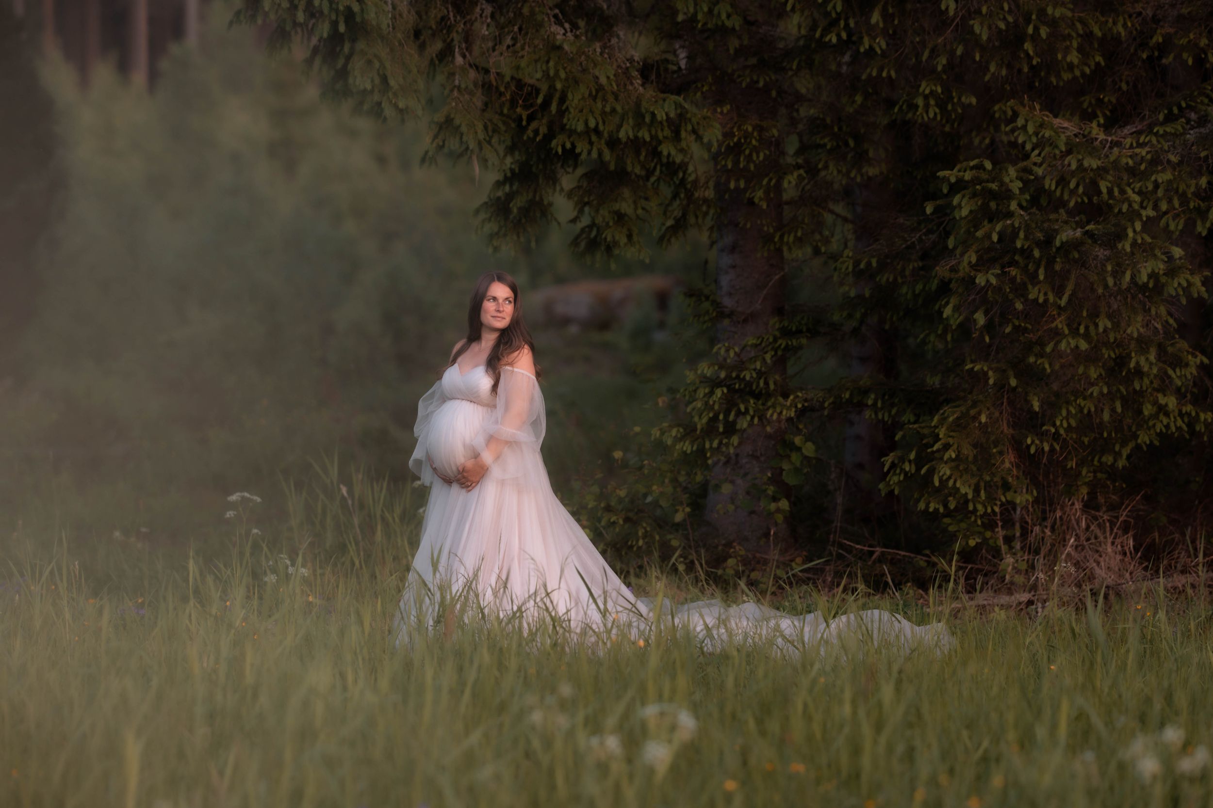 Pregnant woman in white lace gown stands in misty green meadow near tall evergreen trees.