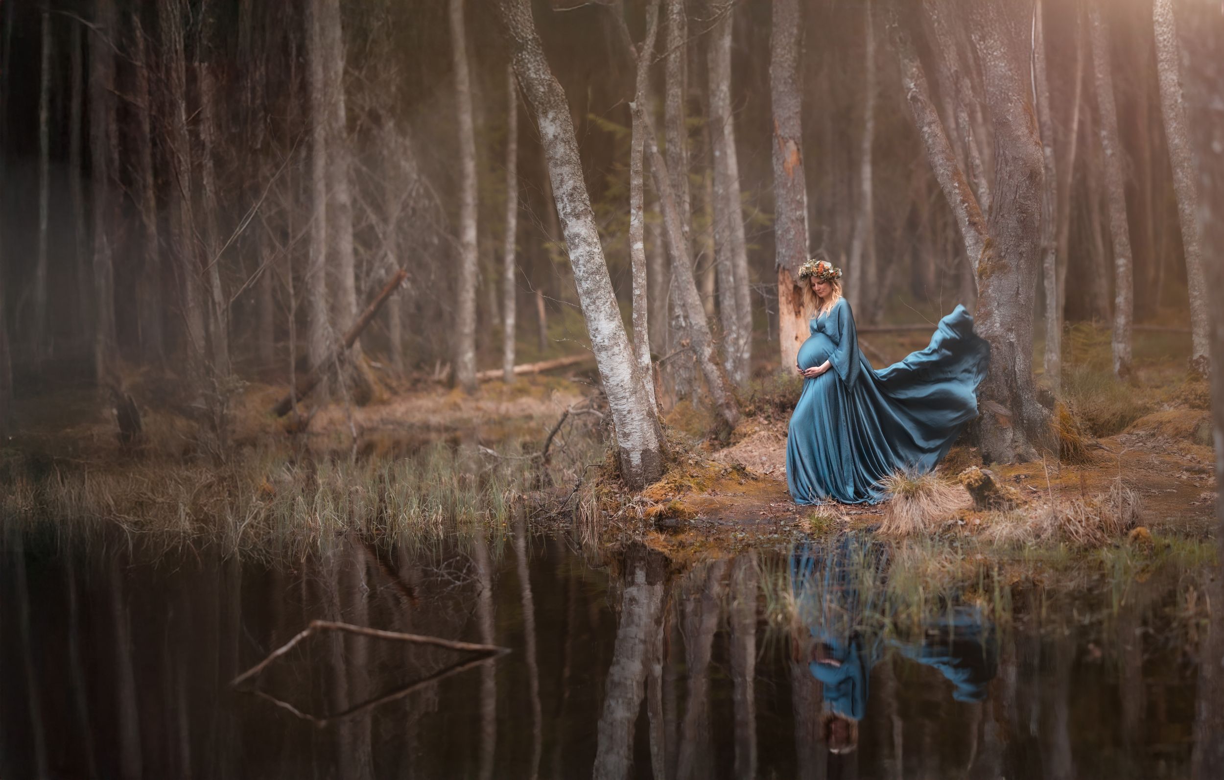 Woman in teal flowing dress stands by reflective forest lake surrounded by birch trees.