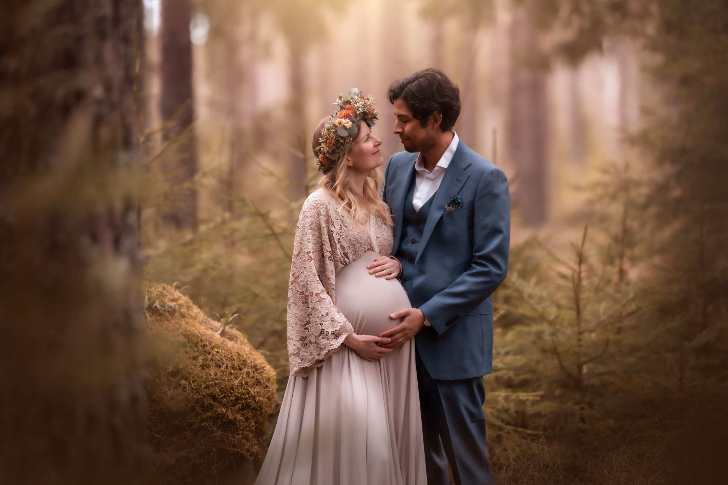 Couple in enchanted forest, pregnant woman in cream gown with floral crown embraced by man in blue suit.