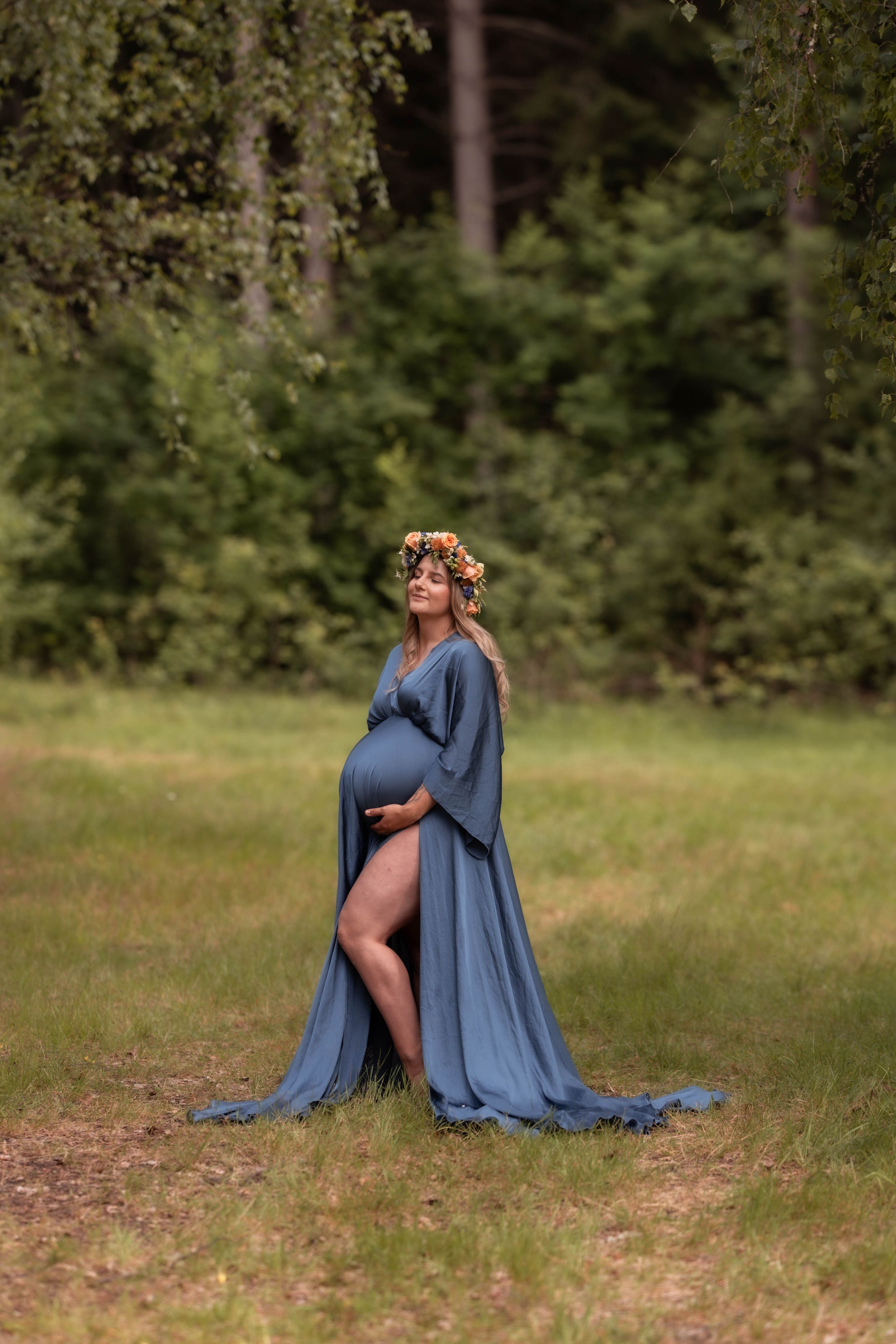 Pregnant woman in blue flowing gown and floral crown stands in green meadow cradling baby bump.