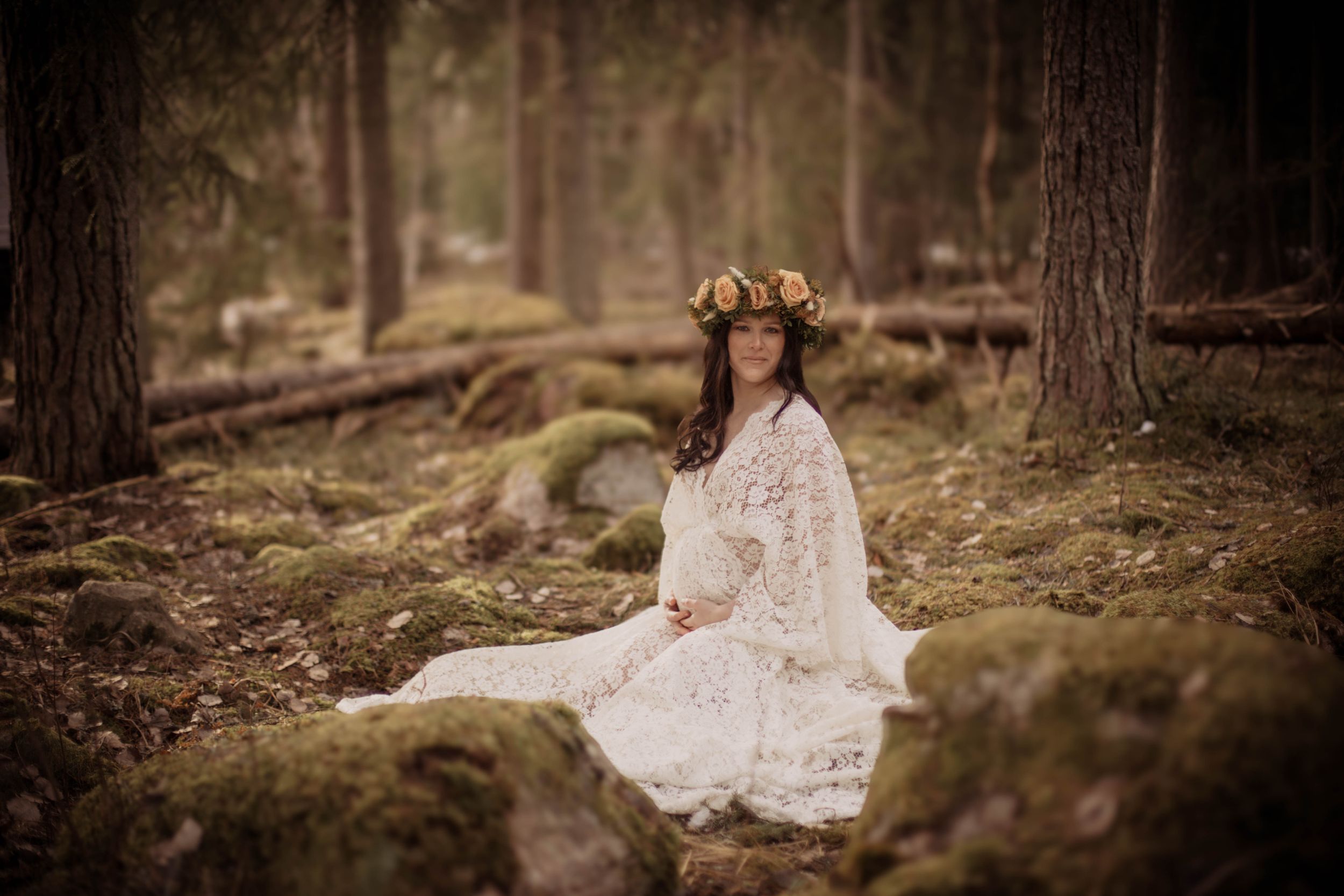 Pregnant woman in white lace dress and floral crown sits peacefully in a mossy forest.