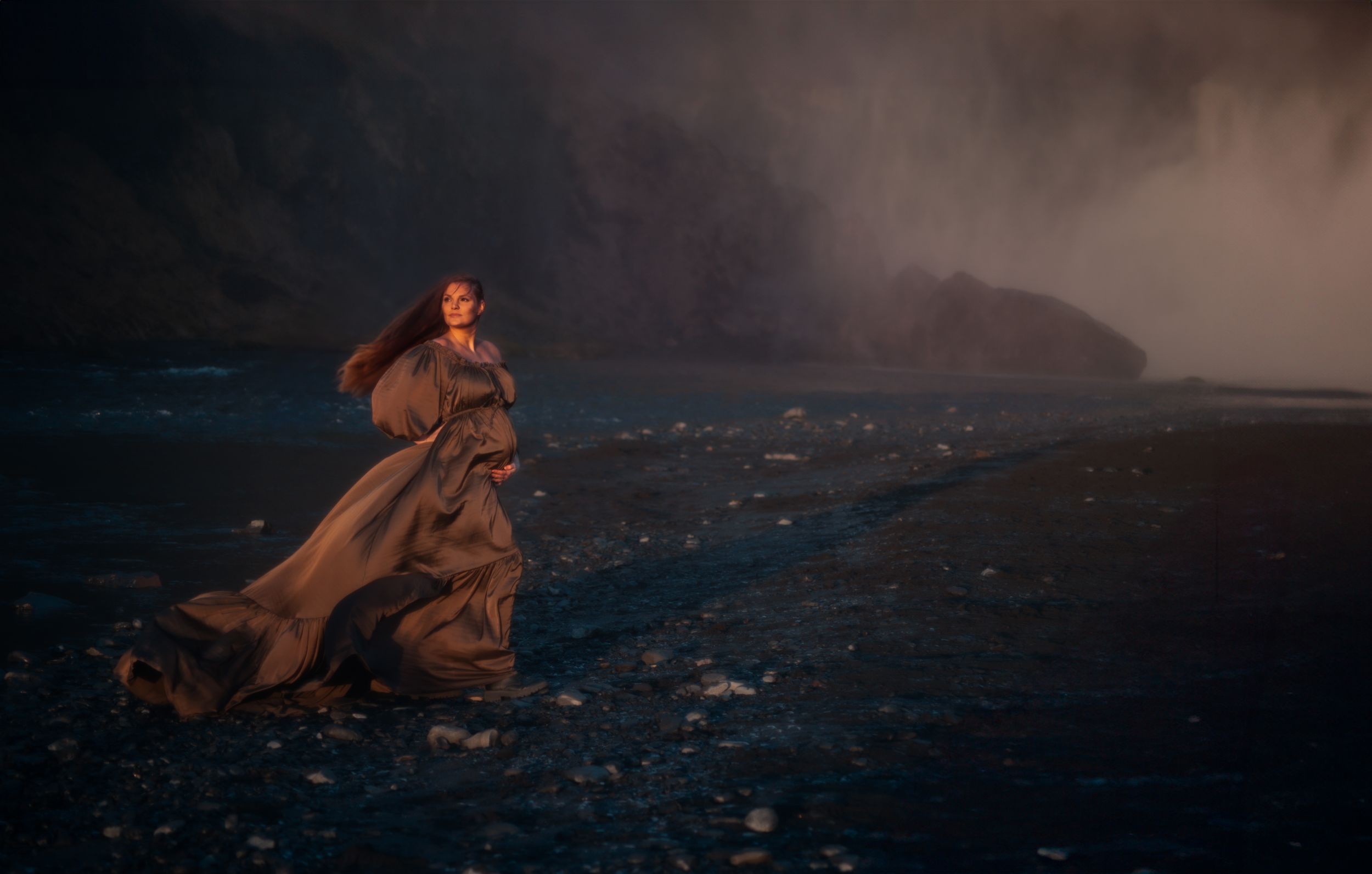 Woman in flowing dark gown stands dramatically on a rocky beach with misty cliffs behind.
