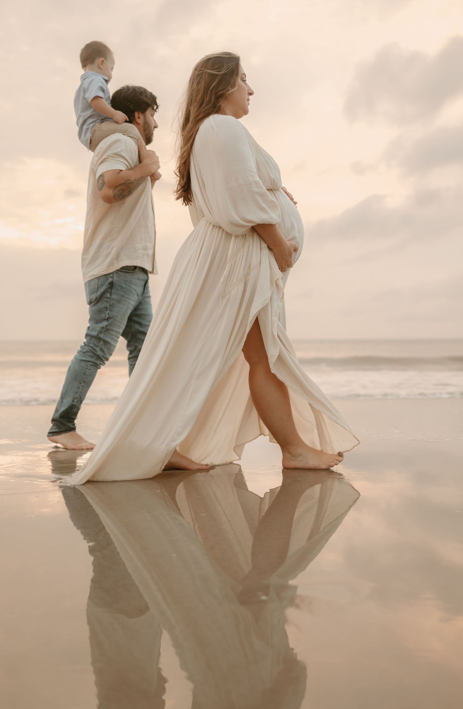 Family in light colored clothing creates reflections while walking along wet beach sand at sunset.