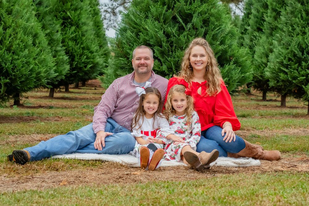 Family poses together at Christmas tree farm on ground surrounded by rows of evergreen trees in winter.