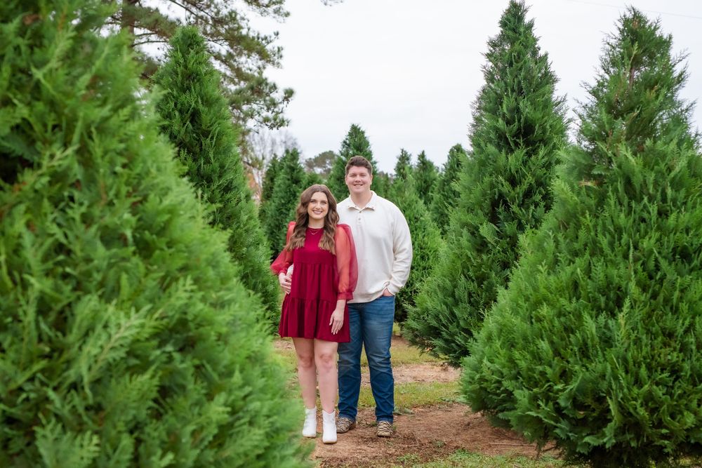 A couple in red and white outfits stands on a dirt path between rows of tall green Christmas trees at a tree farm.