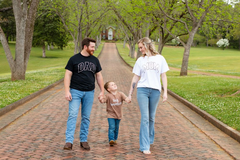 A happy family of three walks hand-in-hand on a brick path lined with green trees, wearing matching Dad and Mom shirts.
