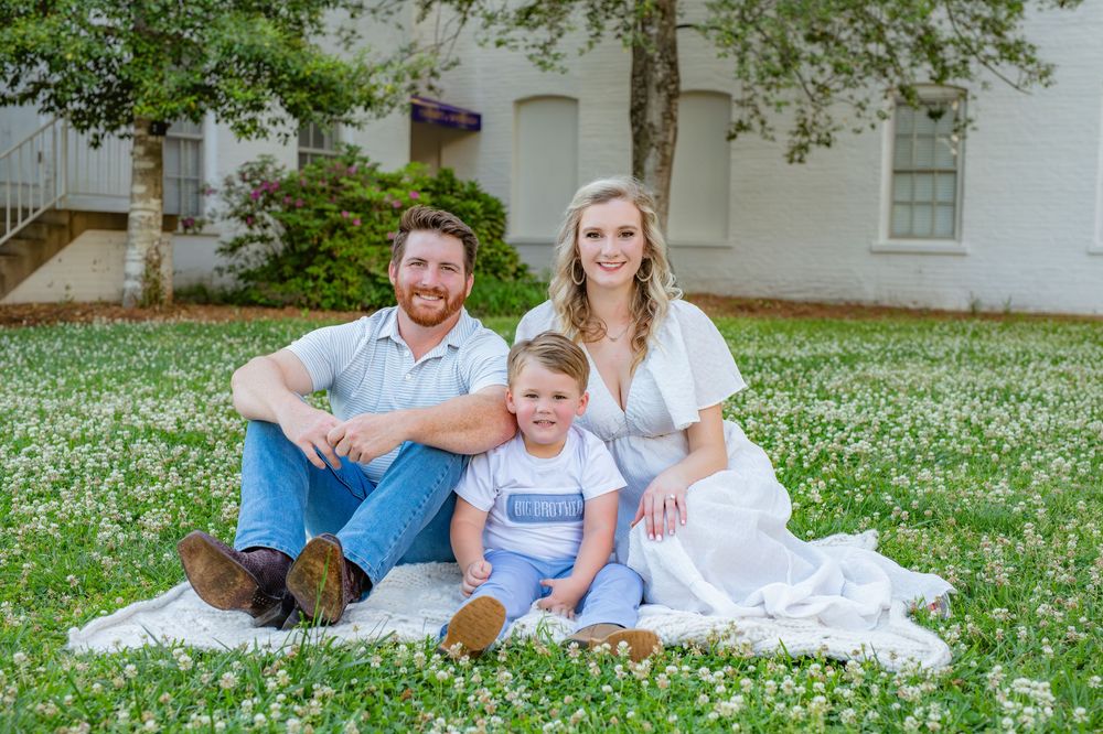 Family of three smiling on a white blanket in a field of daisies, parents flanking a young child outdoors.