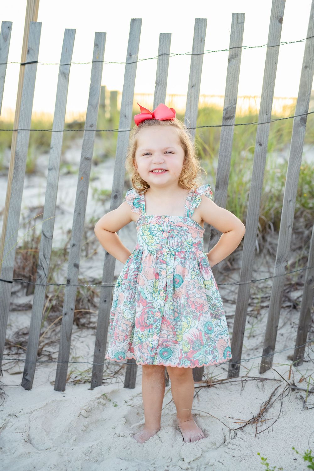 Small child in floral sundress and red bow standing on beach sand by wooden fence at sunset.