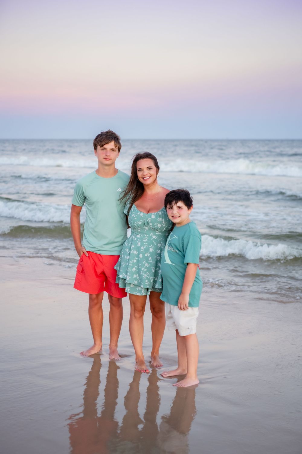 Three people wearing coordinated teal and coral outfits pose together on a beach at sunset with ocean waves in background.