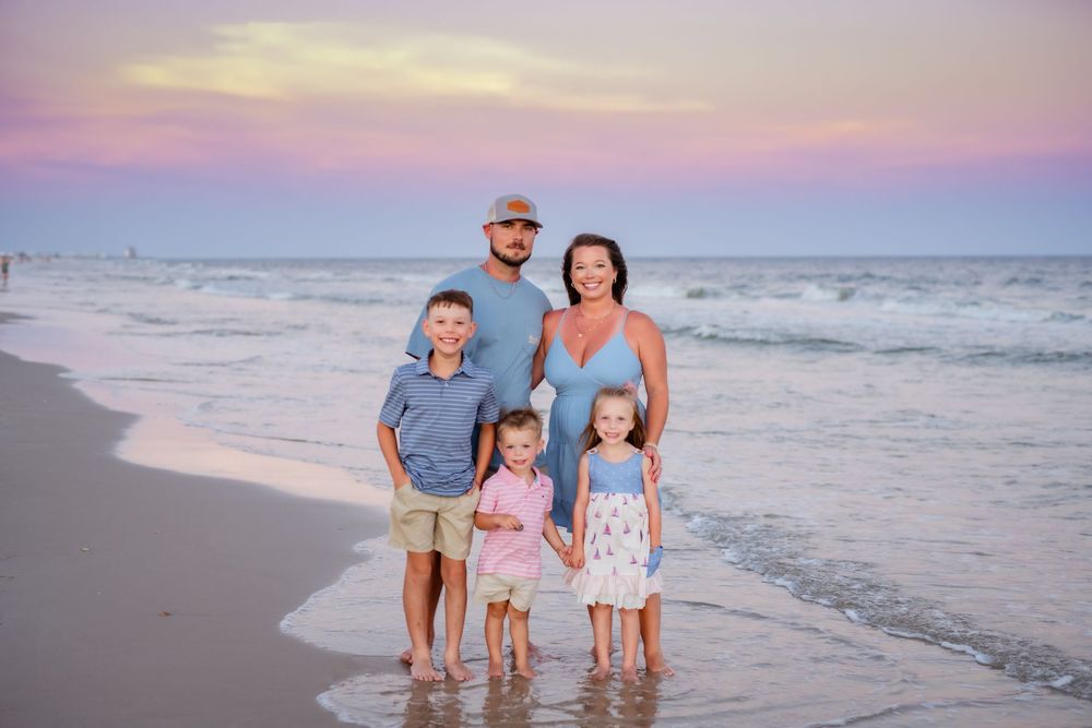 A group poses together at sunset on a beach with gentle waves and pink sky in the background.