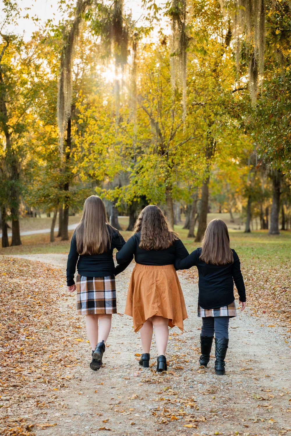 Three people walking together on a path covered in autumn leaves with golden sunlight filtering through trees.