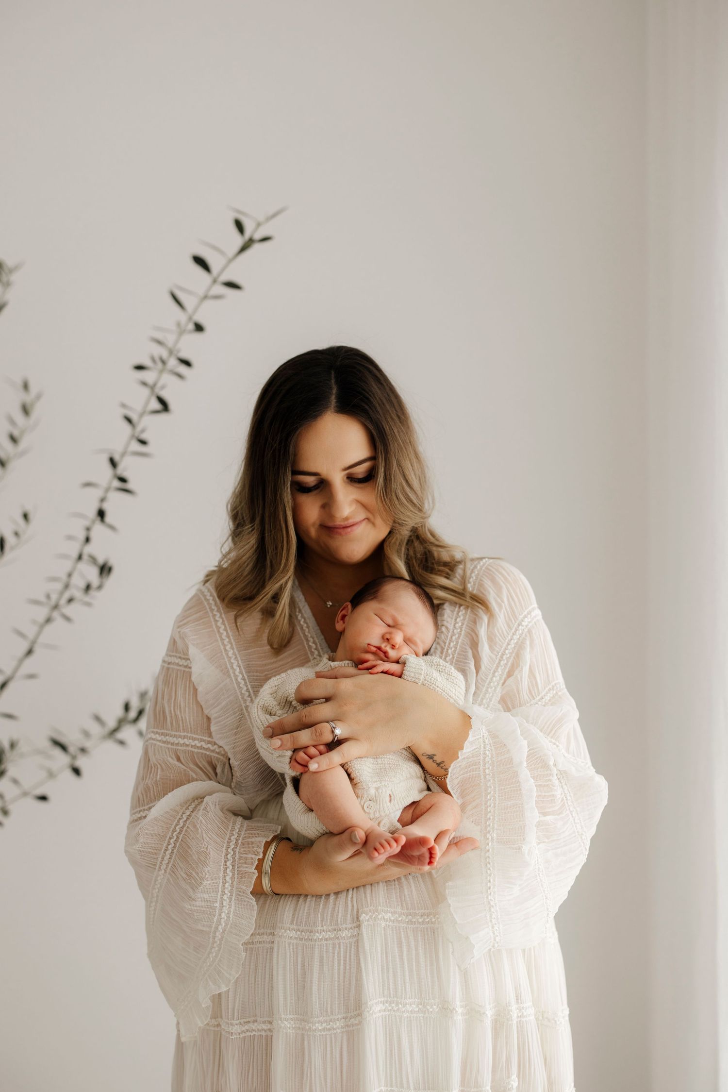 Mother in white boho dress tenderly holds sleeping newborn baby against a bright white background.