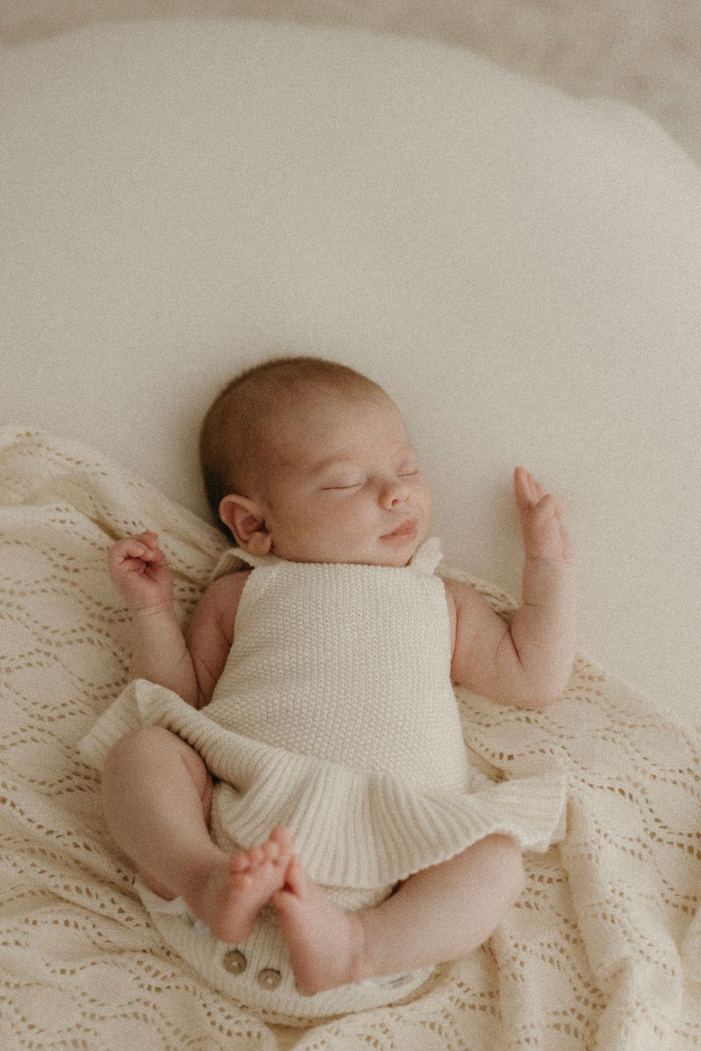 Sleeping newborn baby in cream knit romper lying on soft textured blanket with arms raised.