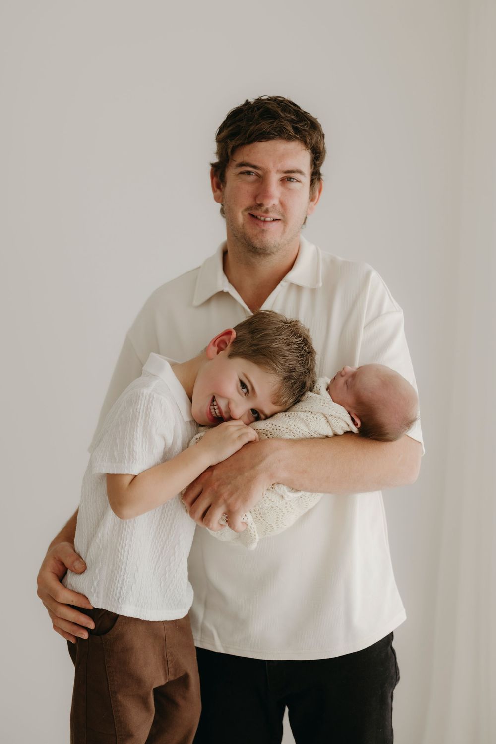 Smiling father in white shirt holds newborn baby while older child hugs him against white background.