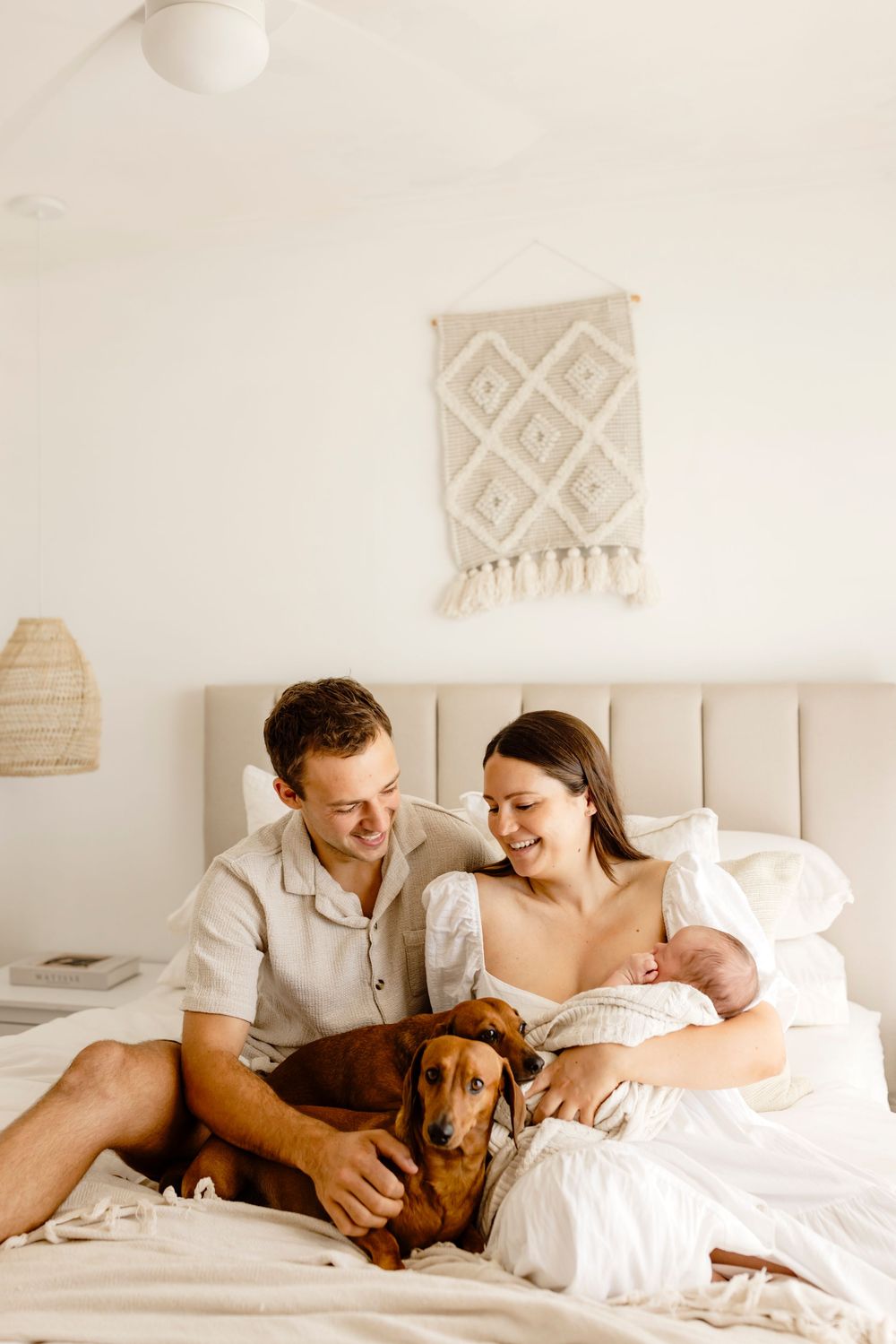 Happy couple sitting on bed with their dachshund dog and newborn baby in bright bedroom.