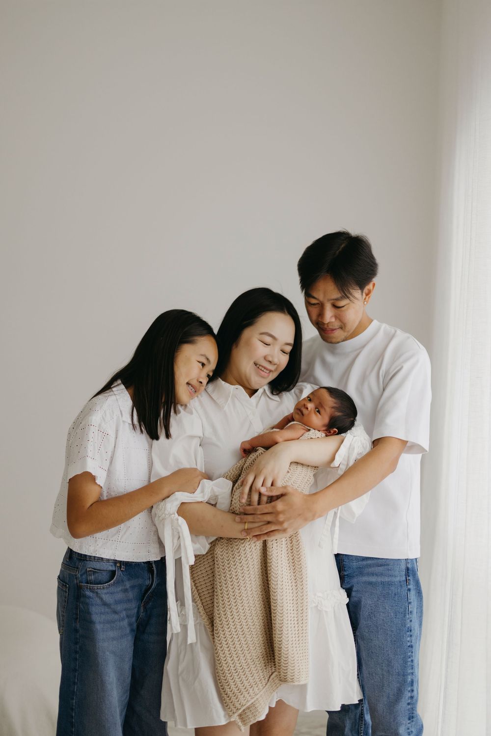 Asian family of four in white tops and jeans lovingly gazing at swaddled newborn baby together.
