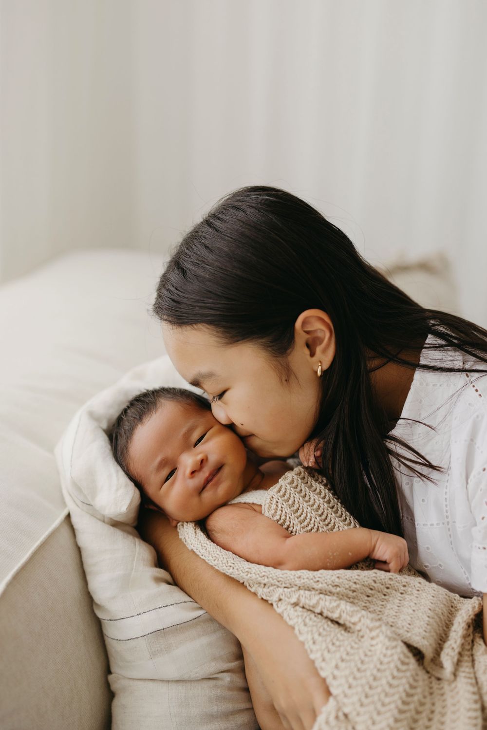 An Asian mother gently kisses her smiling newborn baby wrapped in a soft knit blanket.