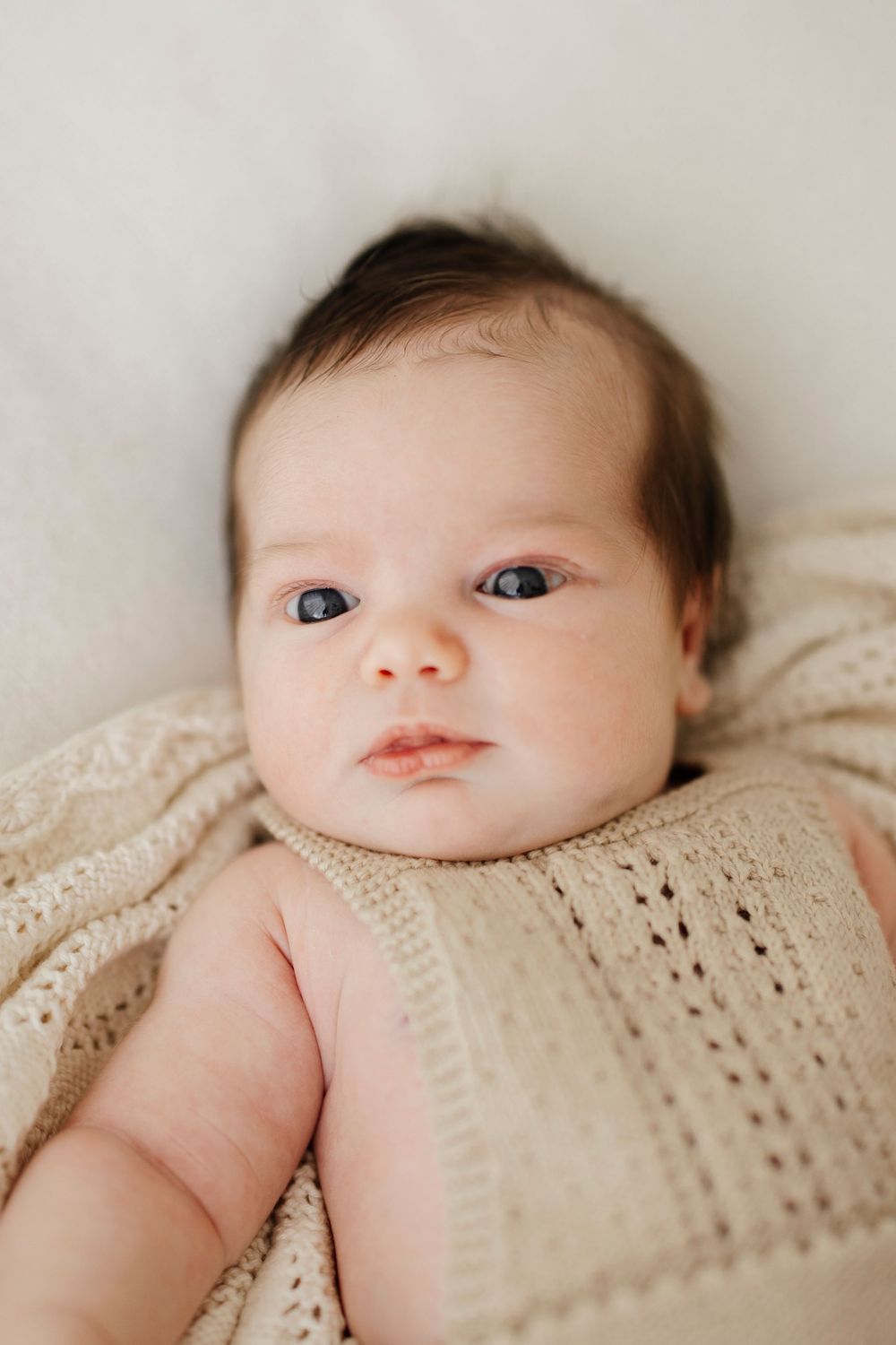 Close-up portrait of an alert newborn baby with dark hair and blue eyes wrapped in a cream knit blanket.