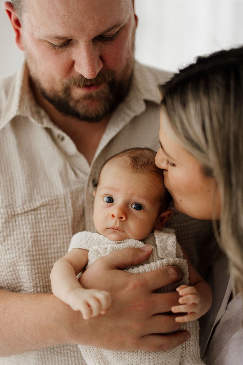 Bearded father and blonde mother lovingly kissing newborn baby with blue eyes.