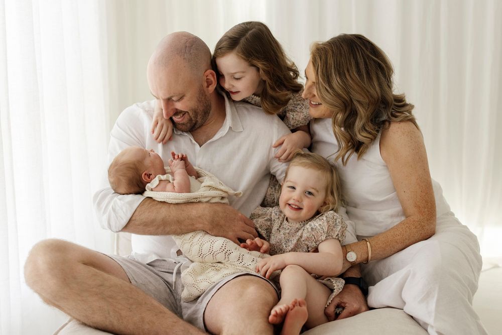 Happy family of five with newborn baby sitting together on white couch.