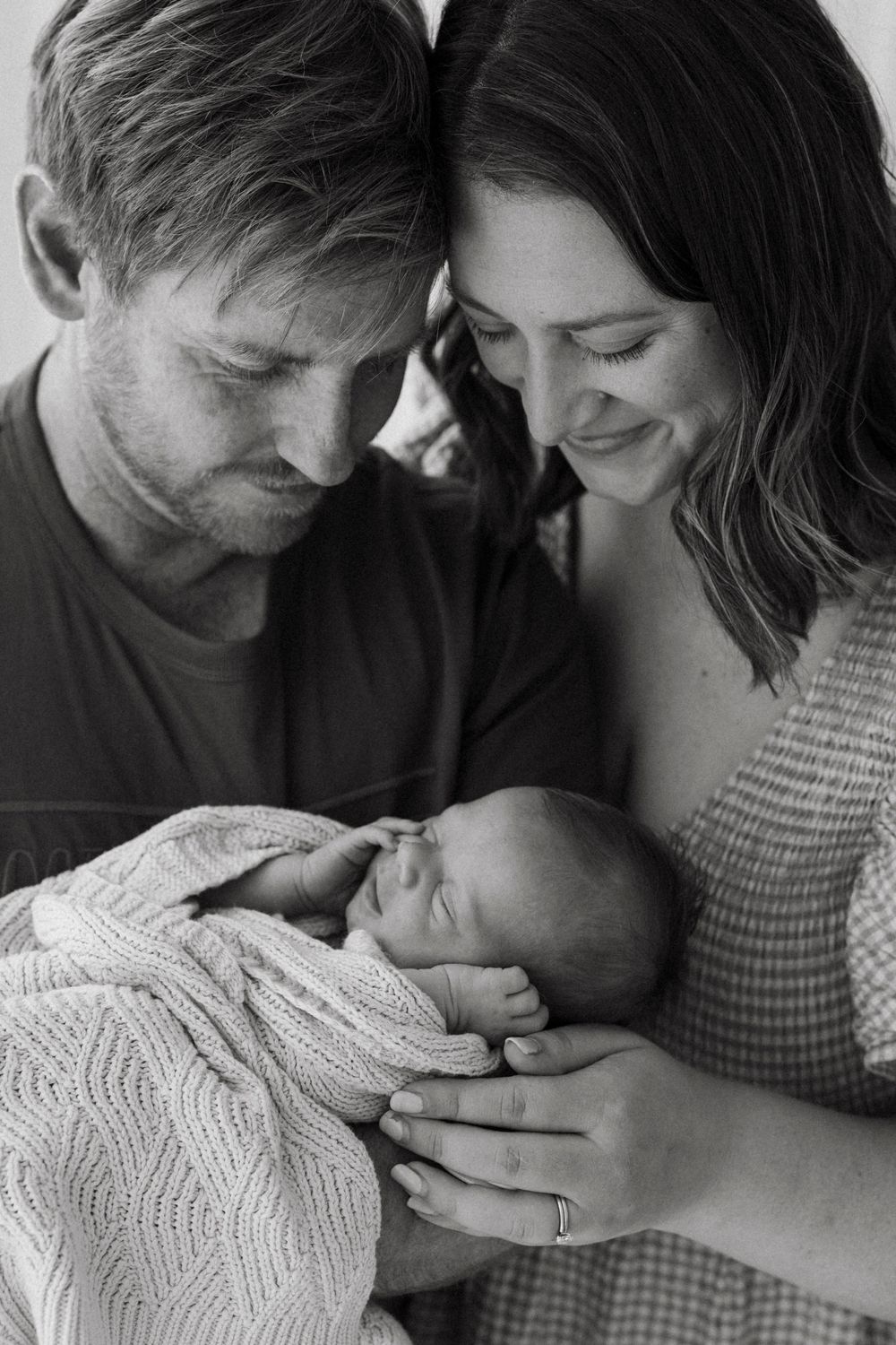 Black and white portrait of parents with foreheads touching lovingly cradling their sleeping newborn.