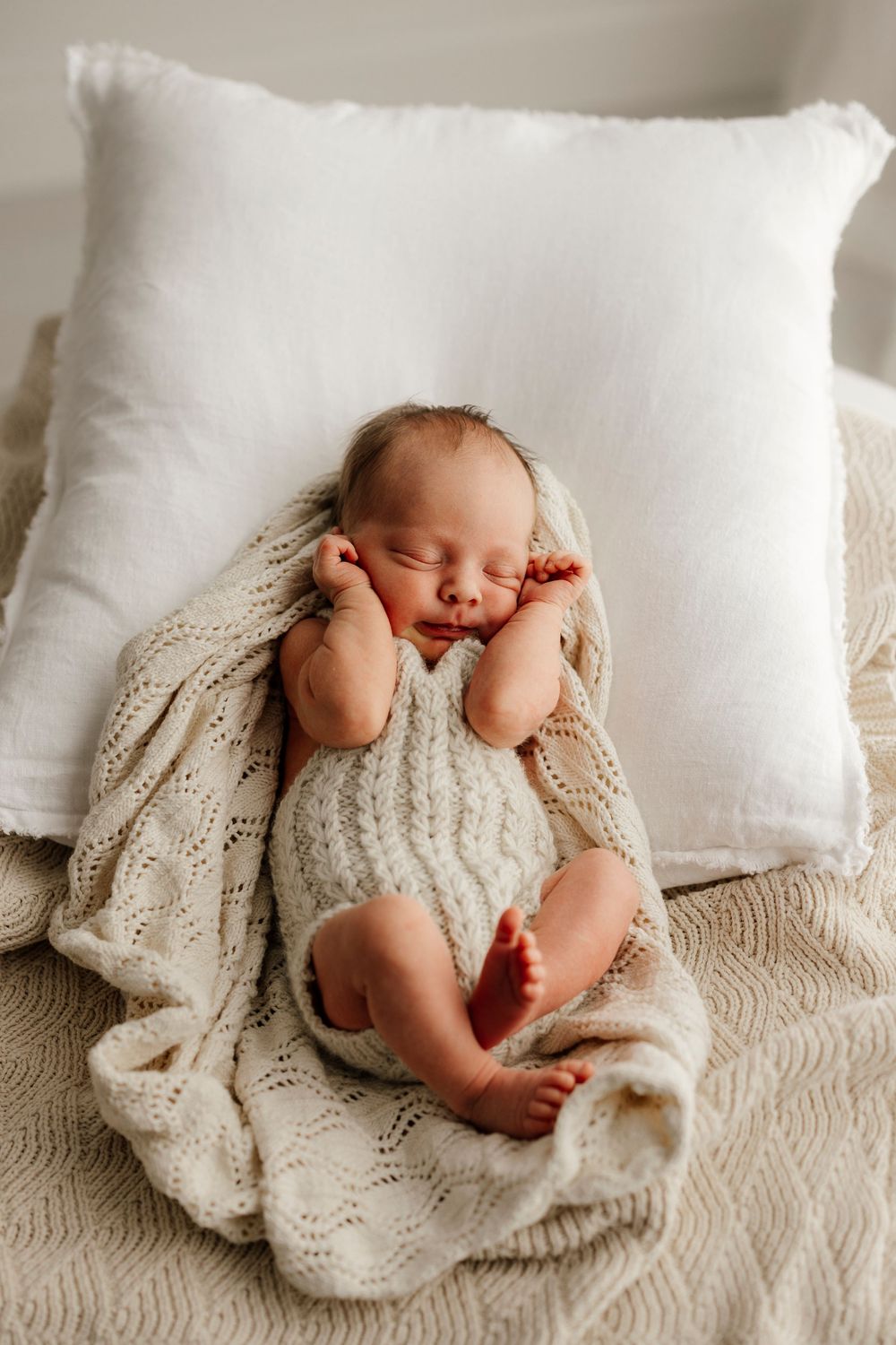 Peaceful newborn baby sleeping in a cream knit romper on a soft blanket beside a white pillow.