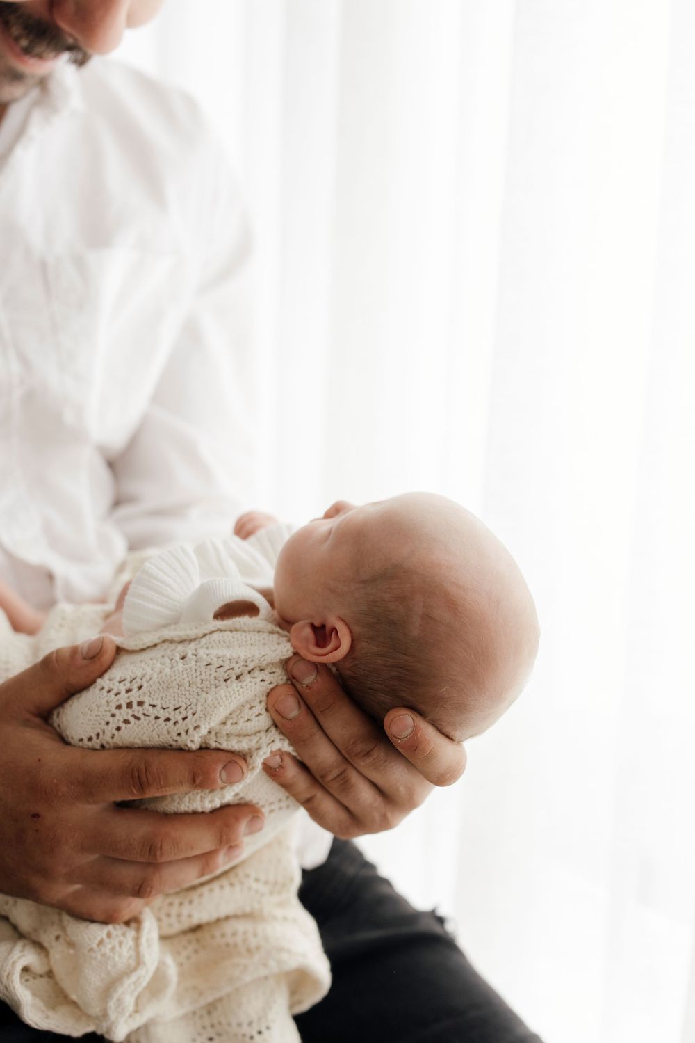 Parent cradling sleeping newborn baby wrapped in cream knit blanket near window.