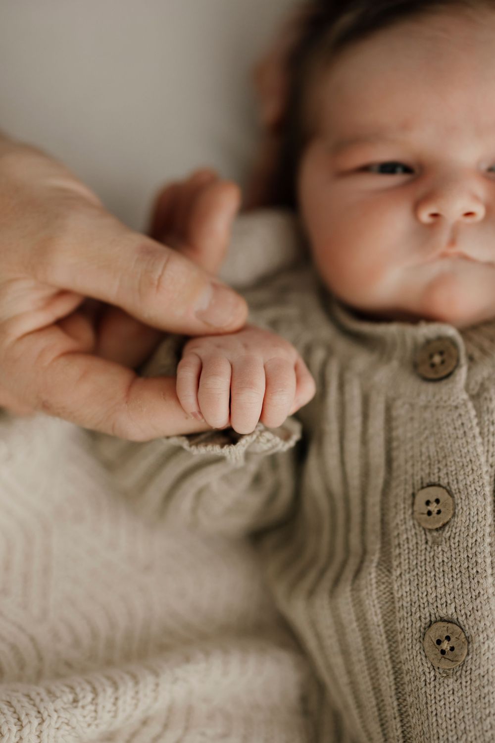 Newborn baby in sage green knit cardigan gently gripping an adult finger.