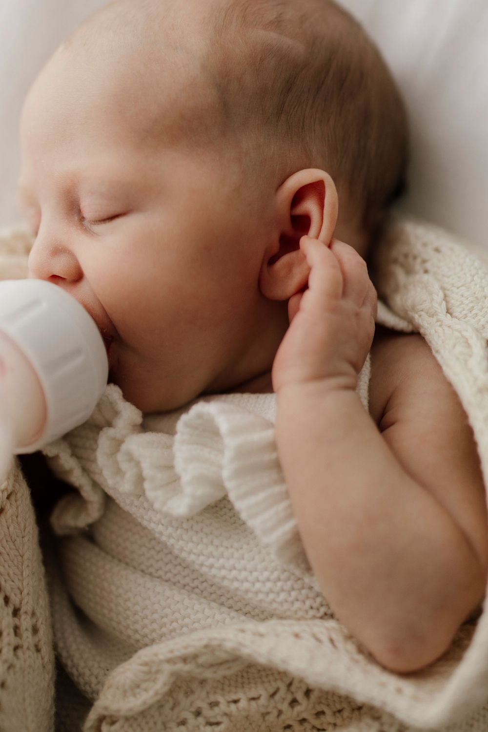 Sleeping newborn baby feeding from bottle wrapped in cream knitted blanket.