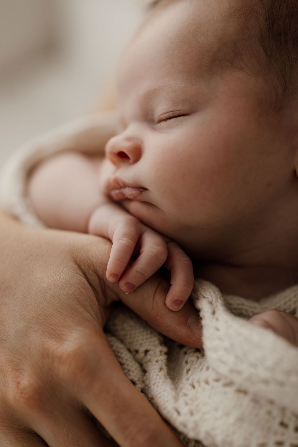 Close-up of sleeping newborn baby with tiny fingers resting on chin wrapped in cream knit blanket.