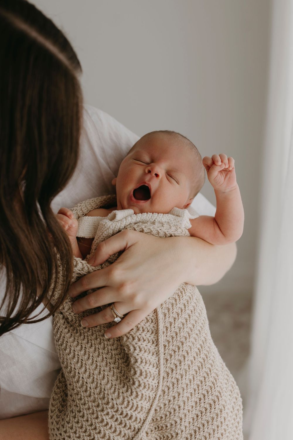 Mother holding yawning stretching newborn baby wrapped in beige knit blanket near bright window.