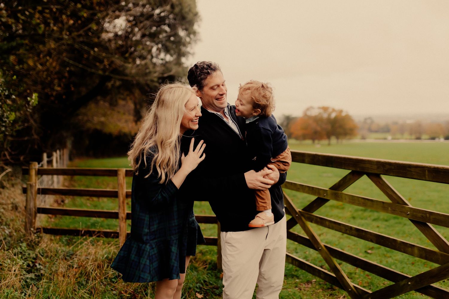 A mum and dad hold and smile at their happy toddler son in a field in front of a wooden fence during a family photo shoot in Chichester