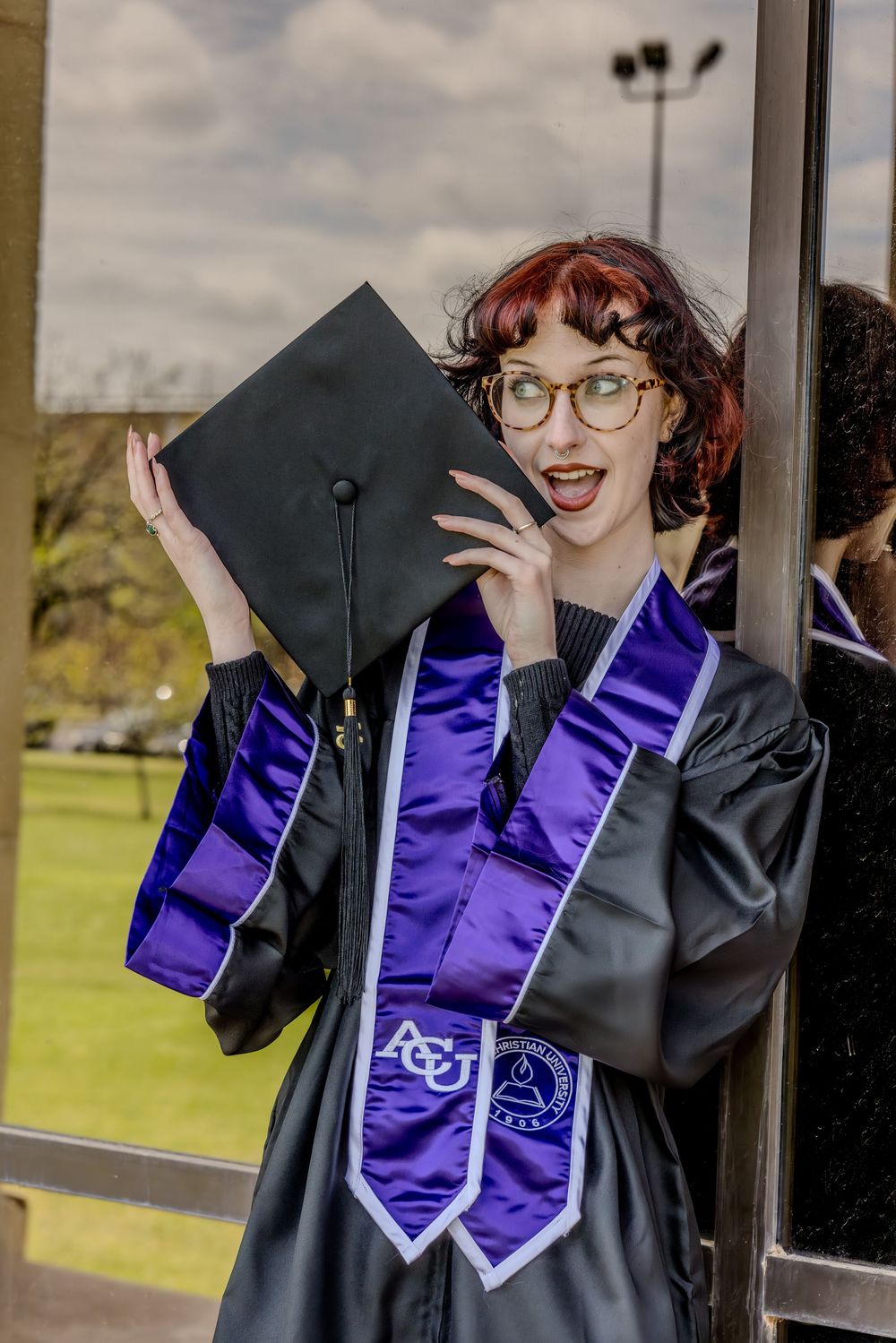 Graduate in black cap and gown with purple stoles celebrates excitedly while holding up graduation cap outdoors.