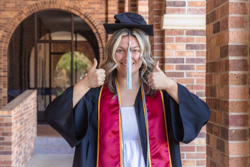 Graduate in red and black graduation regalia gives thumbs up in front of brick archway at university campus.