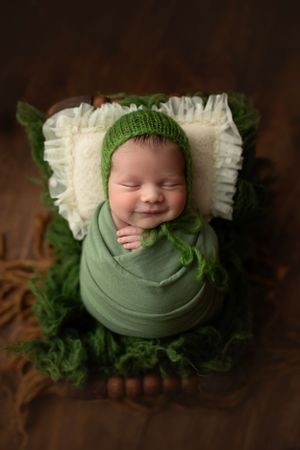 Newborn baby wrapped in green with matching knit hat on white pillow.