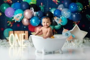 Festive first birthday setup with baby in bathtub surrounded by blue and colorful balloons and glowing ONE sign.