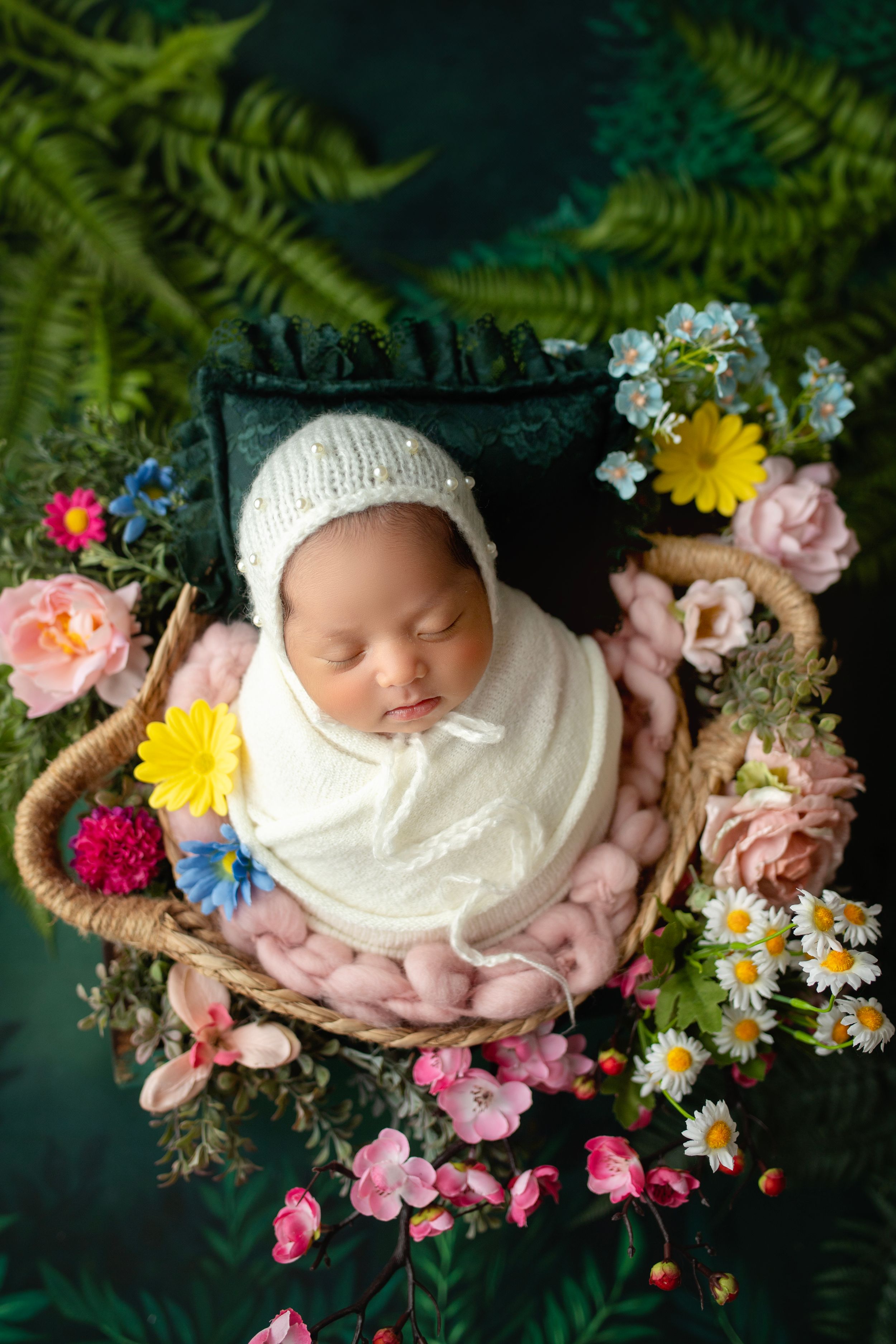Newborn portrait with knit bonnet in basket surrounded by colorful spring flowers and ferns.