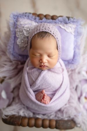 Newborn portrait with lavender wrap and lace bonnet on decorative pillow.