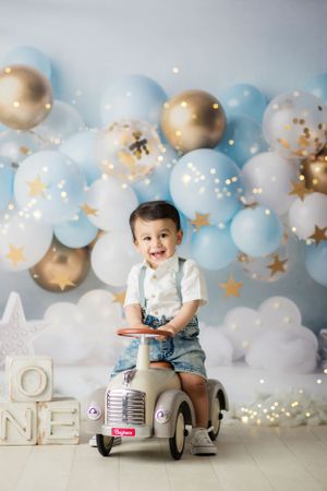 Baby photo session with vintage toy car riding against blue and gold balloon backdrop.