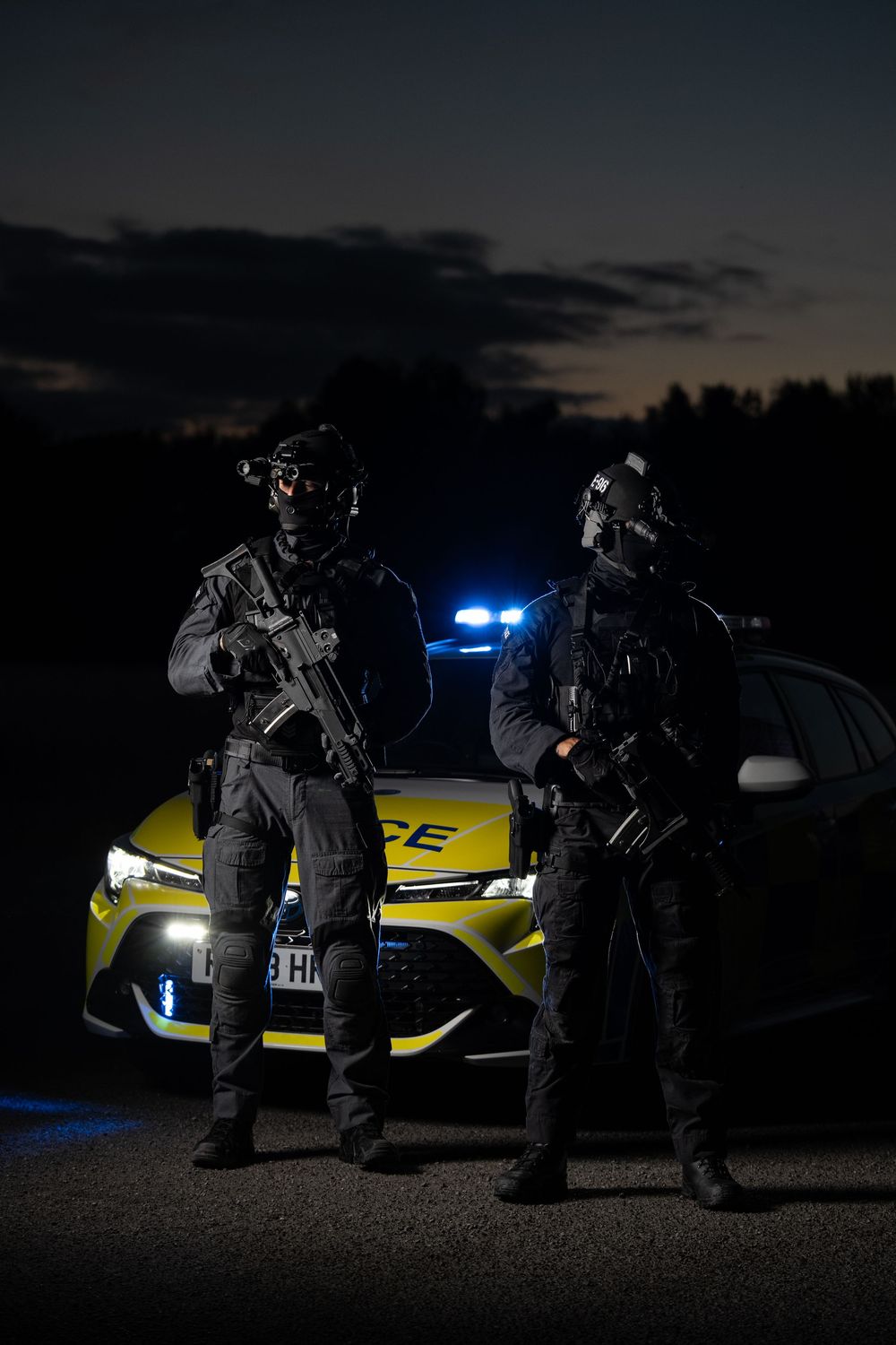 Law enforcement officers conduct a tactical night operation near an illuminated yellow police vehicle in low light conditions.