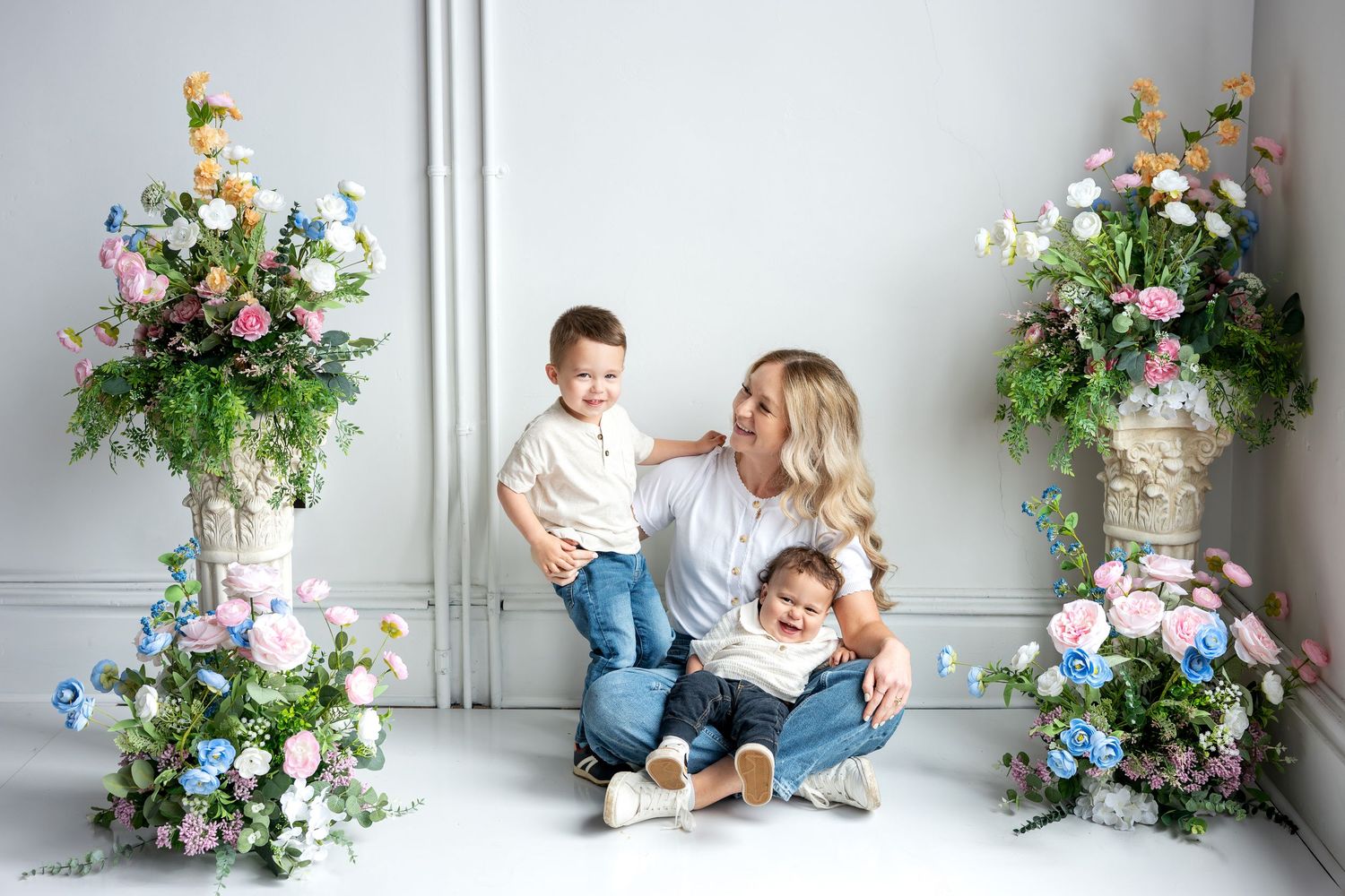 A woman sits on the floor with two young children surrounded by colorful floral arrangements in a bright studio.