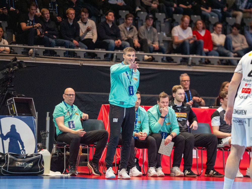 Handball players in white and turquoise uniforms compete during an indoor match with spectators in the background.