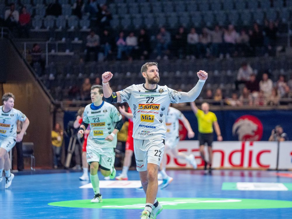 Handball players compete in an intense match on a blue court in a professional indoor arena.